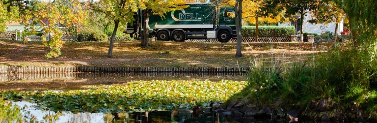 KELL-Müllfahrzeug im Hintergrund zwischen Bäumen; im Vordergrund ein Teich mit Seerosen und herbstlicher Vegetation.