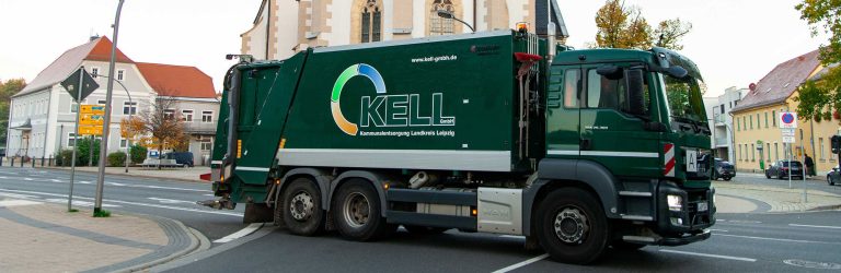 Grünes KELL-Müllfahrzeug auf einer Straße im Ort; im Hintergrund Gebäude und eine Kirche.